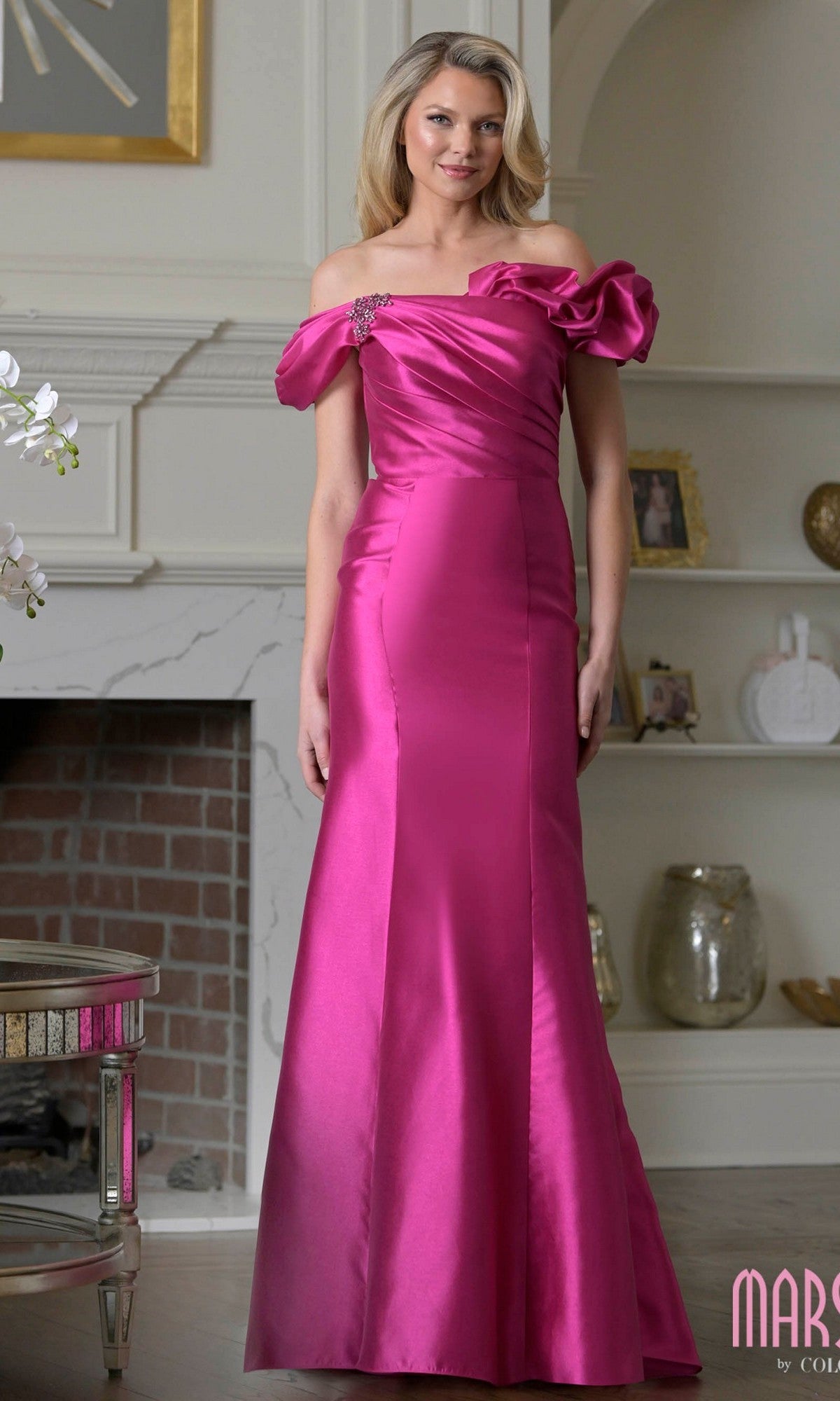 A woman stands indoors wearing the Marsoni MV1407 long formal dress in fuchsia, featuring ruffles, an off-shoulder design, floor-length satin fabric, and a decorative shoulder detail.
